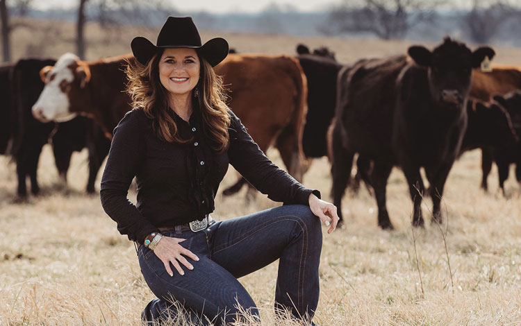 Woman with black hair wearing a black hat and kneeling in a pasture with cattle behind her.